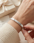 A close-up of a silver engraved bracelet being adjusted on a person's wrist, with a cream ribbed sweater sleeve visible and a soft gift box in the background.