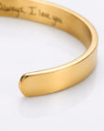 A close-up of a polished gold cuff bracelet with engraved script text inside, shown on a clean white background.