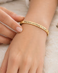 A gold-toned metal cuff bracelet engraved with the words 'Today tomorrow & Always, I love you' worn on a person's wrist, with their other hand gently resting nearby.