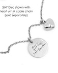 Close-up of a necklace featuring a circular disc with an engraved message and a heart-shaped urn, attached to a cable chain.