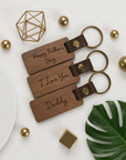 Wooden keychains with engraved messages on a decorative background with gold elements and a green leaf.