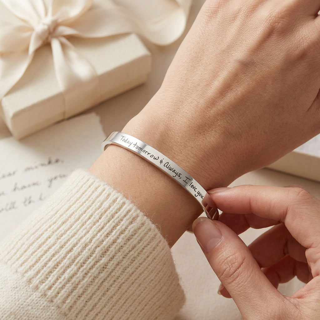 A close-up of a silver engraved bracelet being adjusted on a person's wrist, with a cream ribbed sweater sleeve visible and a soft gift box in the background.