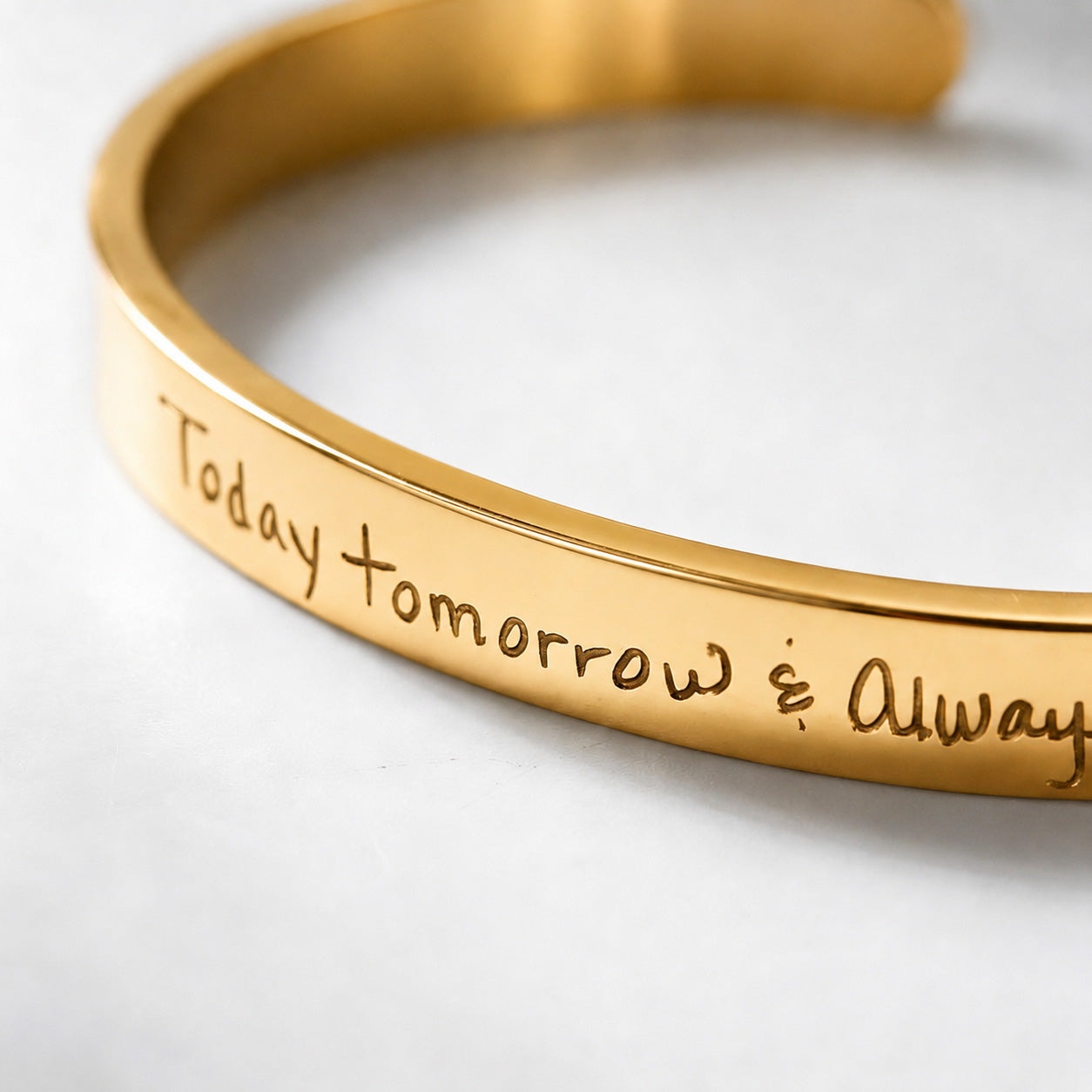 A close-up of a gold metal cuff bracelet engraved with the phrase 'Today tomorrow &amp; Always' in a handwritten font, resting on a light gray surface.