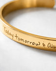 A close-up of a gold metal cuff bracelet engraved with the phrase 'Today tomorrow & Always' in a handwritten font, resting on a light gray surface.