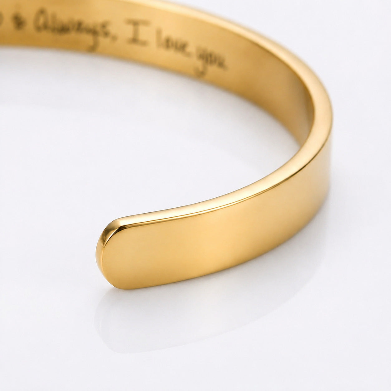 A close-up of a polished gold cuff bracelet with engraved script text inside, shown on a clean white background.