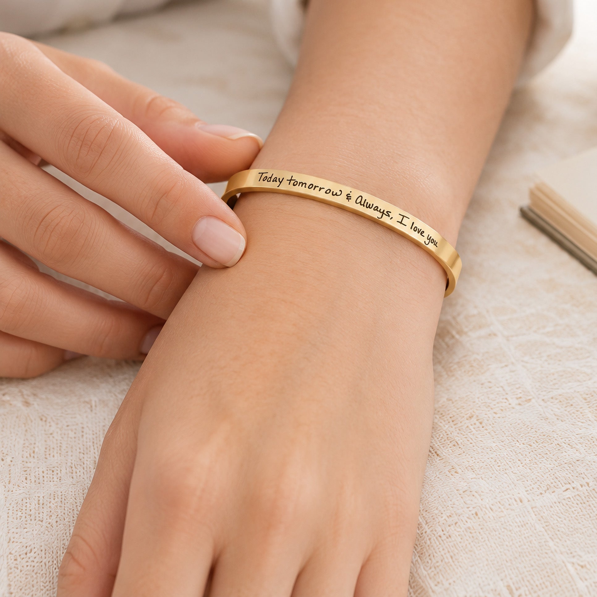A gold-toned metal cuff bracelet engraved with the words 'Today tomorrow &amp; Always, I love you' worn on a person's wrist, with their other hand gently resting nearby.