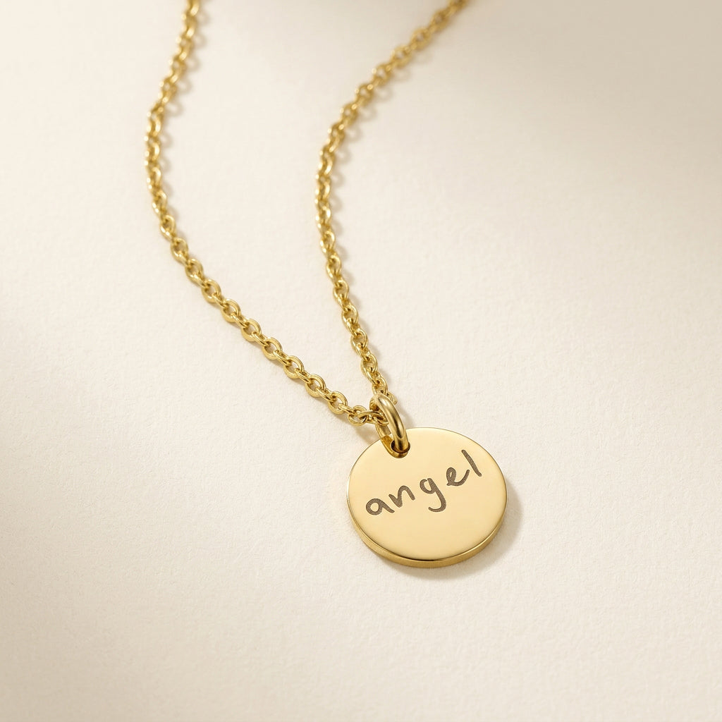 Gold necklace with a round pendant inscribed with the word 'angel' on a beige background