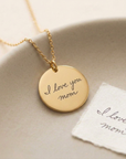 Gold necklace with 'I love you mom' pendant on a beige surface