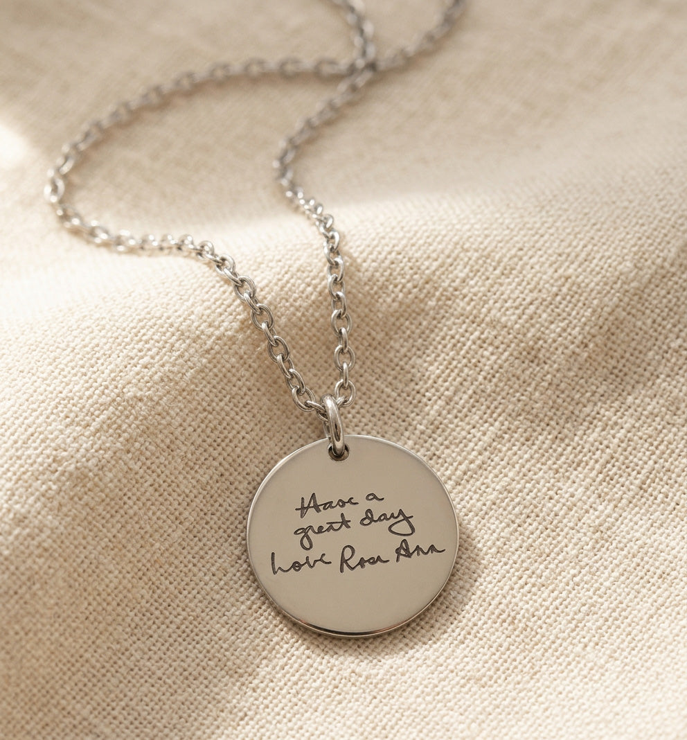 A silver round disc pendant necklace with the engraved text 'Have a great day here, Rom Ant' resting on a textured beige linen fabric background.