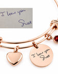 Rose gold double-band expandable bracelet with a heart charm, a round disc charm engraved with 'I love you Shot', a small red gemstone charm, and a small inset showing a handwritten note that reads 'I love you Shot'.