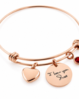 Rose gold bangle bracelet with a heart charm, a round disc charm engraved with 'I love you Scott', and a small red gemstone accent, shown on a white background.