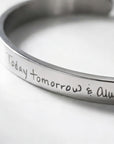 A close-up of a polished silver cuff bracelet engraved with the handwritten phrase 'Today tomorrow & Always'.