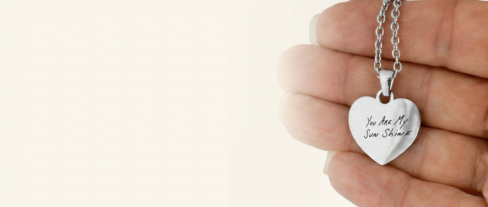 A silver heart pendant necklace engraved with 'You Are My Sun Shine' held in a person's hand against a warm off-white background.
